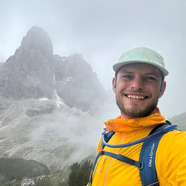 Selfie van een jongen met een pet en gele jas voor een bergtop